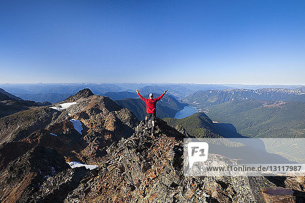 Rückansicht eines Wanderers mit erhobenen Armen auf einem Berg stehend vor klarem Himmel