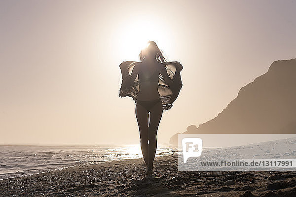 Frau in voller Länge im Bikini  während sie bei Sonnenuntergang am Strand Textilien gegen den klaren Himmel hält