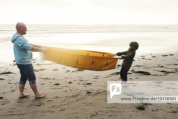 Seitenansicht eines spielerischen Vaters und eines Sohnes  die an einer Picknickdecke ziehen  während sie am Strand vor klarem Himmel am Ufer stehen