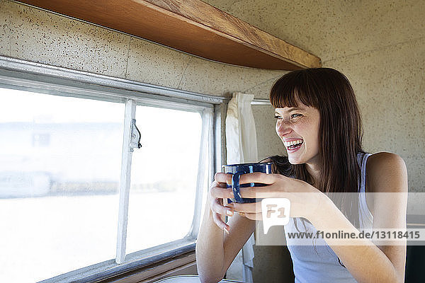 Fröhliche Frau trinkt Kaffee  während sie im Wohnmobil am Fenster sitzt