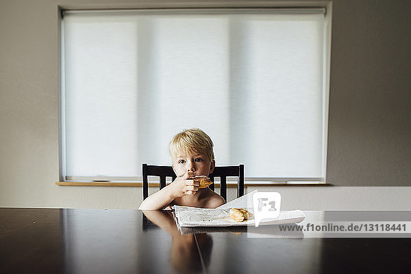 Portrait of shirtless boy eating bread while sitting by table at home