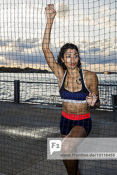 Nachdenkliche Sportlerin schaut weg  während sie bei Sonnenuntergang am Netz gegen den Fluss steht