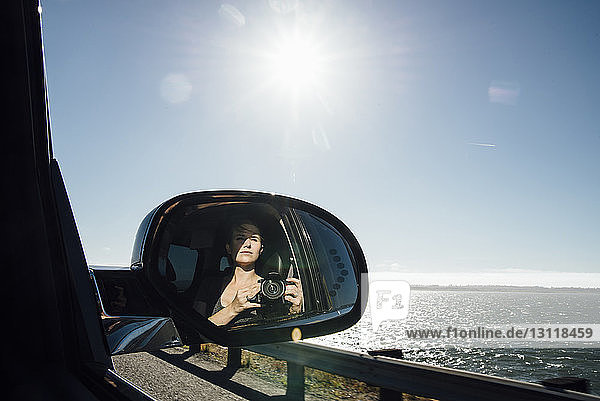 Woman photographing while reflecting on side-view mirror of car against sky