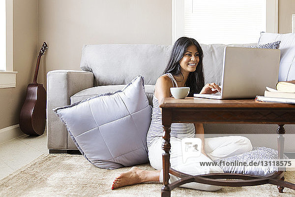 Glückliche schwangere Frau surft mit Laptop am Tisch im Haus im Internet
