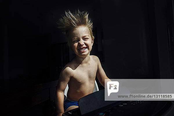 Portrait of happy shirtless boy enjoying breeze from air conditioner in darkroom at home