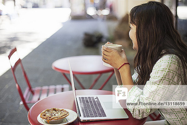 Seitenansicht einer nachdenklichen Frau mit Kaffeebecher in der Hand während sie mit ihrem Laptop im Straßencafé sitzt