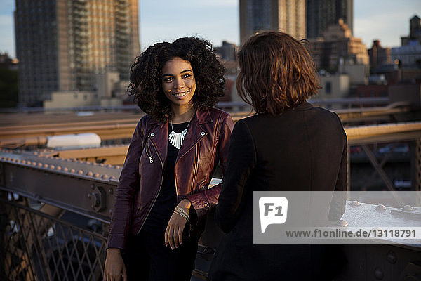 Freundinnen unterhalten sich  während sie bei Sonnenuntergang am Geländer der Brooklyn Bridge stehen