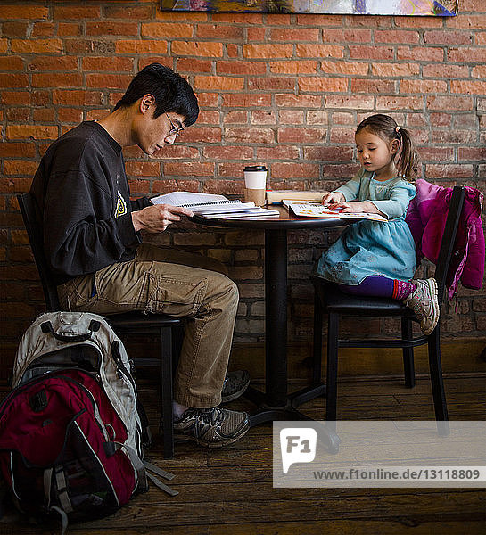 Father working while daughter reading book while sitting against brick wall at cafe