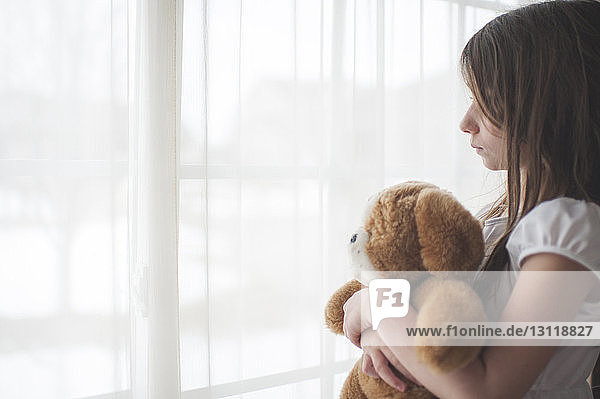Mädchen hält Teddybär und schaut zu Hause durchs Fenster