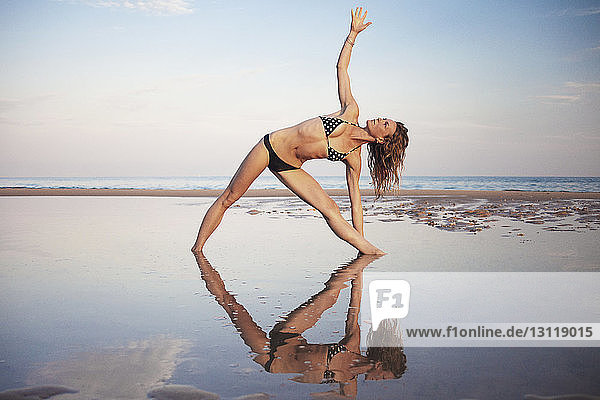Frau im Bikini beim Yoga am Strand gegen den Himmel