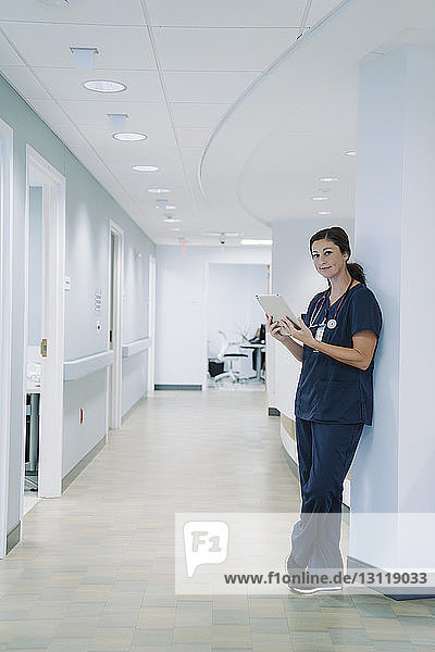 Porträt einer Ärztin  die einen Tablet-Computer benutzt  während sie sich im Krankenhaus an eine architektonische Säule lehnt