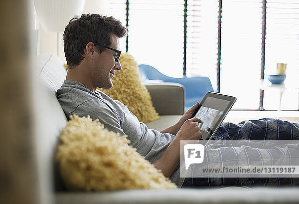 Lächelnder Mann benutzt Tablette  während er sich zu Hause auf dem Sofa ausruht