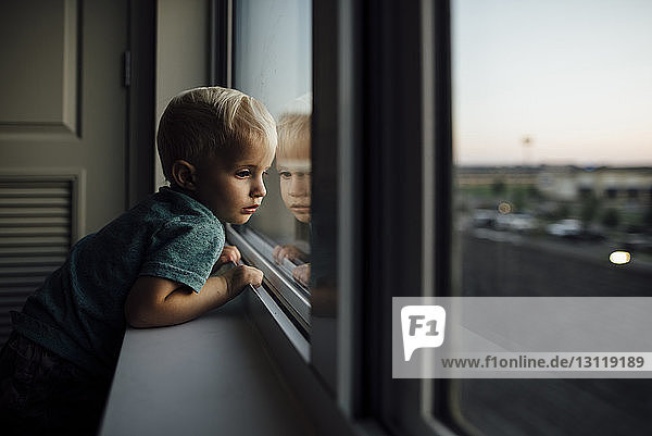 Seitenansicht eines kleinen Jungen  der zu Hause stehend durch ein Fenster schaut