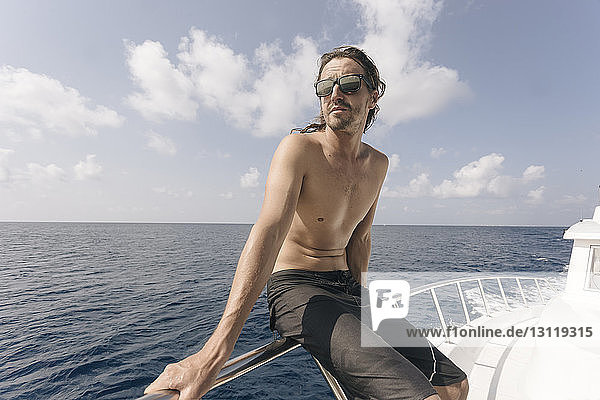 Mann ohne Hemd mit Sonnenbrille  während er auf einer Jacht in der Reling auf dem Meer gegen den Himmel sitzt