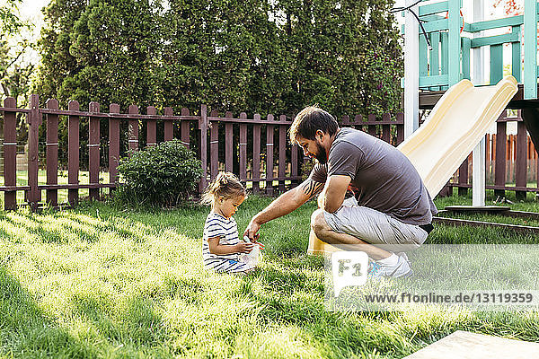 Father playing with daughter sitting on grassy field at backyard