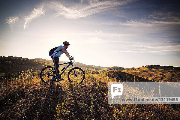 Männlicher Athlet auf Fahrrad in grasbewachsenem Feld vor bewölktem Himmel