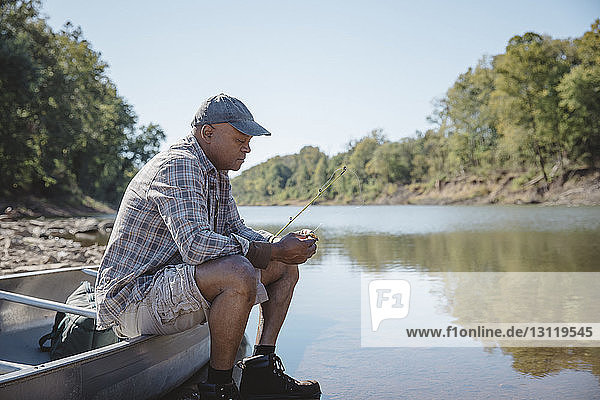 Man adjusting fishing tackle while sitting on boat by lake against clear sky