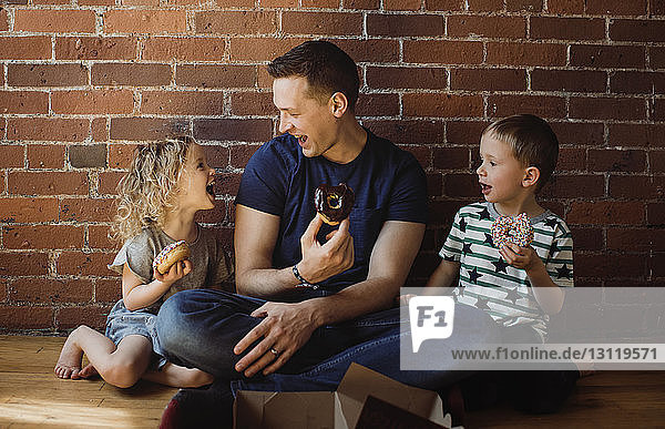 Glücklicher Vater mit Kindern  die Donuts essen  während sie zu Hause auf dem Boden sitzen