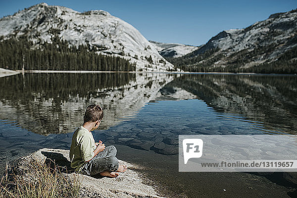 Side view of boy sitting on rock by lake against mountains at Yosemite National Park