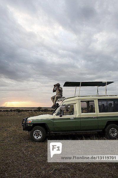 Mann schaut durch ein Fernglas  während er auf einem Geländewagen im Serengeti-Nationalpark sitzt
