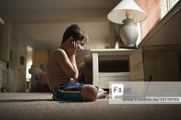 Seitenansicht eines aufgeregten Jungen  der einen Laptop-Computer betrachtet  während er auf einem Teppich sitzt