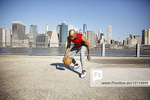 Ein Mann in voller Länge spielt mit Basketball auf der Promenade gegen die Skyline der Stadt