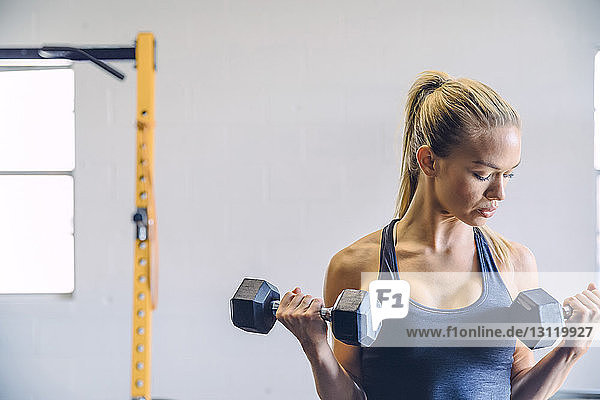 Young woman lifting dumbbells while exercising against wall in gym