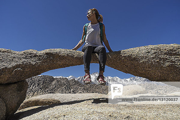 Low angle view of female hiker sitting on natural arch against clear sky