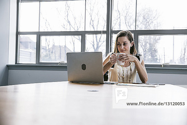 Geschäftsfrau hält Getränk in der Hand und schaut auf Laptop-Computer  während sie im Sitzungssaal sitzt