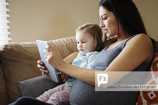 Nahaufnahme einer schwangeren Frau  die der Tochter die Tablette zeigt  während sie zu Hause auf dem Sofa sitzt