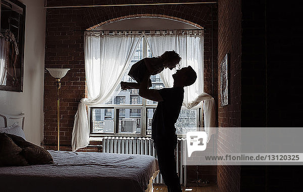Silhouette father picking up daughter while standing by bed at home