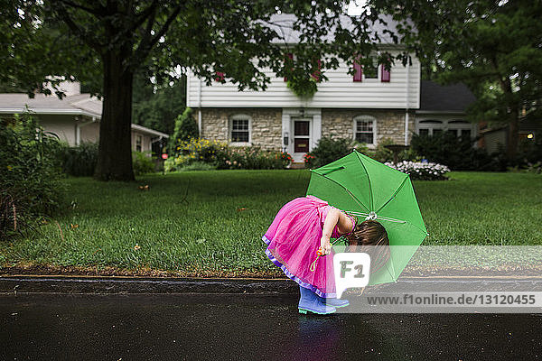 Seitenansicht eines Mädchens mit grünem Regenschirm in rosa Kleid beim Bücken auf nasser Straße