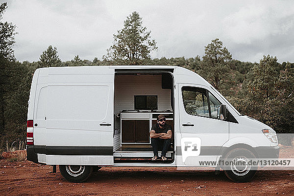 Man with arms crossed sitting in motor home against cloudy sky at forest