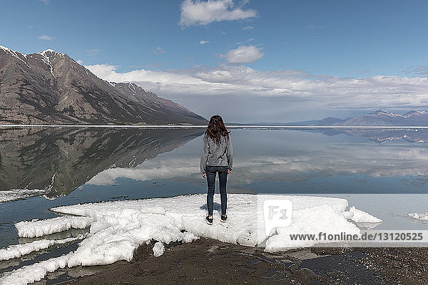 Woman standing on snow by river while looking at view against mountains and sky