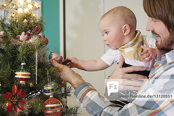 Vater zeigt dem Baby zu Hause in der Nähe des Weihnachtsbaums Ornament