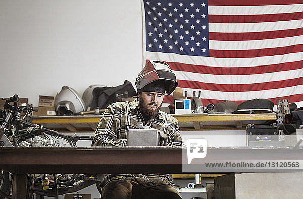 Serious manual worker working on machinery against American flag at auto repair shop