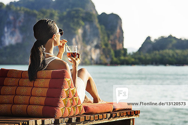 Frau genießt Getränk  während sie auf Liegestuhl gegen das Meer sitzt