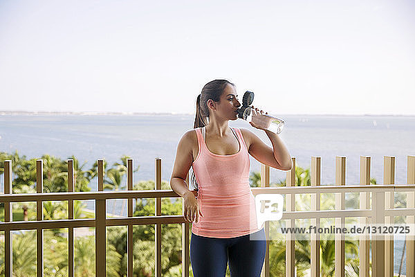 Woman drinking water while standing at railing against sea