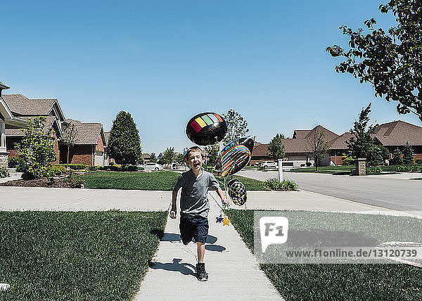 Glücklicher Junge mit Luftballons  der bei strahlend blauem Himmel am sonnigen Tag auf dem Fußweg läuft