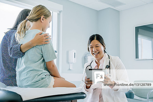 Cheerful pediatrician looking at tablet computer while mother and daughter sitting on examination table