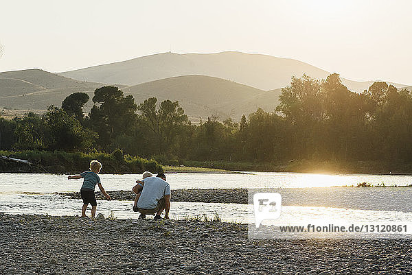 Rückansicht eines Vaters mit Söhnen beim Spielen am Seeufer vor klarem Himmel bei Sonnenuntergang