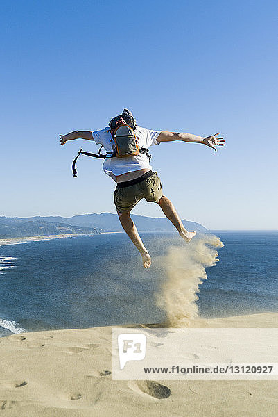 Rückansicht eines verspielten Mannes  der am Strand vor strahlend blauem Himmel auf Sand springt