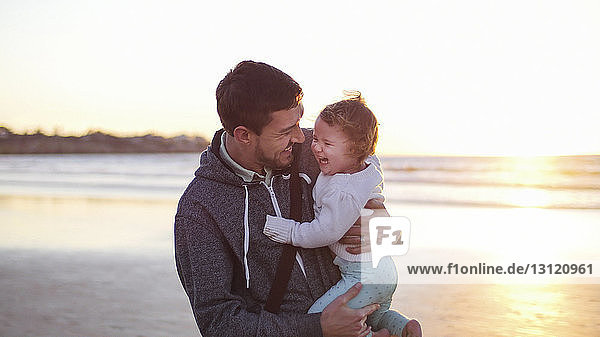 Happy father carrying daughter while standing at beach against clear sky during sunset