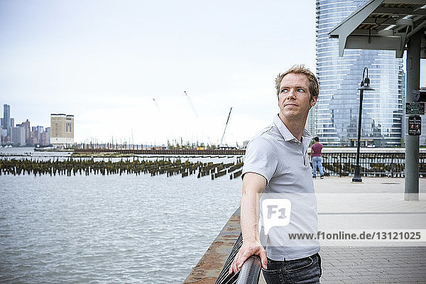 Side view of thoughtful man looking away while standing by river on promenade against sky in city