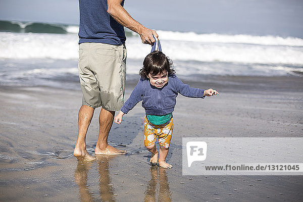 Low section of playful father pulling daughter's jacket while standing on shore at beach