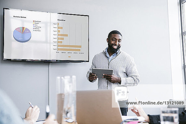 Lächelnder Geschäftsmann hält Tablet-Computer in der Hand  der bei einer Besprechung im Sitzungssaal Grafiken erklärt