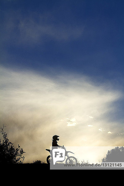 Silhouette of motocross racer against cloudy sky during sunset