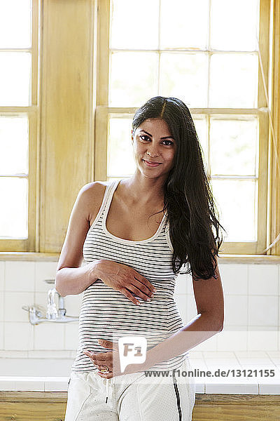 Smiling pregnant woman standing against window at home