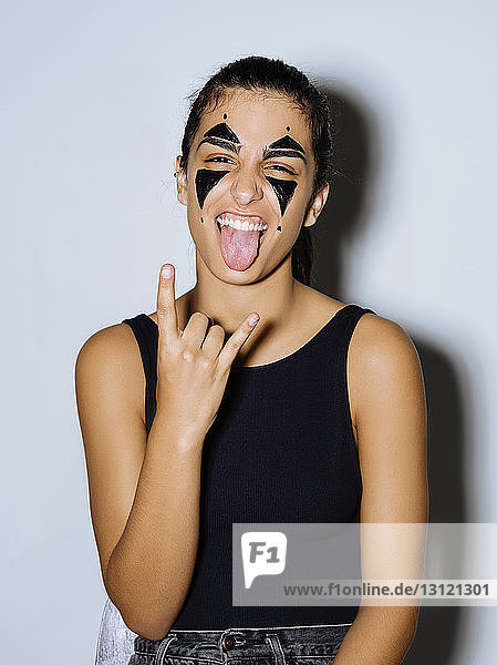 Close-up portrait of happy woman with face paint sticking out tongue while standing against wall