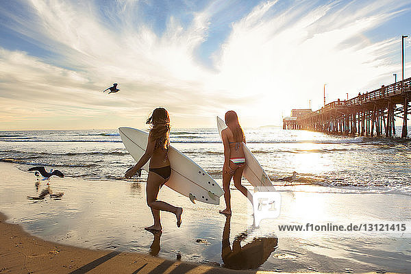 Female friends holding surfboards and walking on wet shore